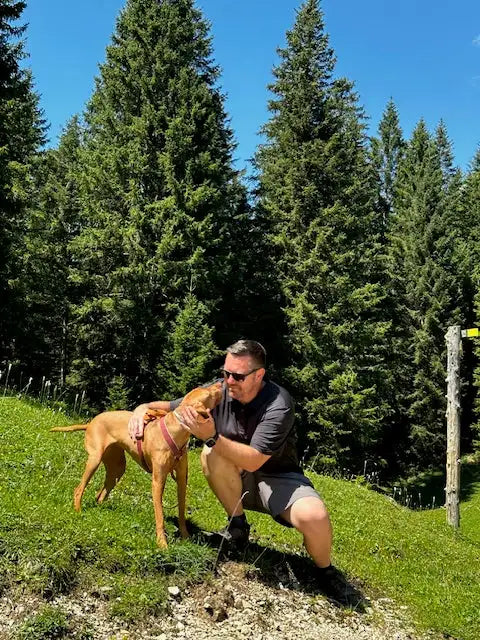 Ein Mann in einem dunkelgrauen Hemd und Shorts kniet auf dem Gras neben einem tanfarbenen Hund mit rotem Halsband, vor dem Hintergrund hoher grüner Kiefern unter einem klaren blauen Himmel.