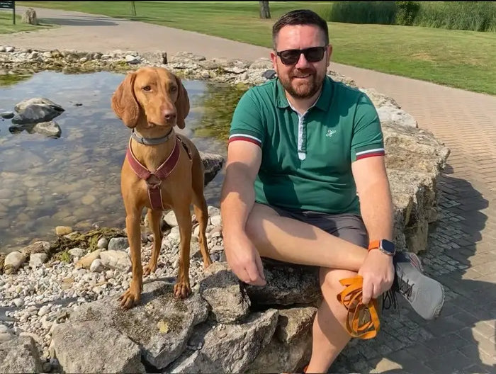 Ein Mann, der ein grünes Poloshirt mit rot- und weißem Besatz an den Ärmeln trägt, sitzt neben einem tanfarbenen Vizsla-Hund, beide positioniert in der Nähe von Felsen an einem flachen Teich.